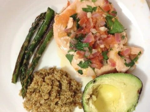 A white plate with Pico de Gallo salmon topped with chopped tomatoes and herbs, alongside grilled asparagus, a serving of quinoa, and half an avocado. The text Pico De Gallo Salmon is in cursive at the top.