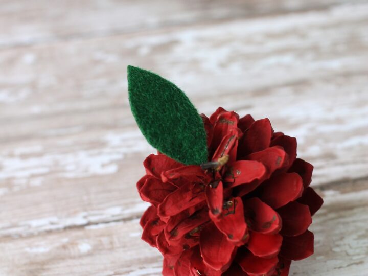A decorative red pine cone painted to resemble an apple, complete with a piece of green felt shaped like a leaf, set against a rustic wooden background.