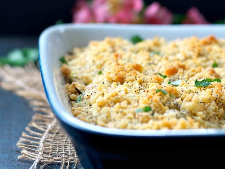 A close-up of a rectangular casserole dish filled with a golden-brown baked dish topped with breadcrumbs and herbs. The dish sits on a textured cloth, with pink flowers blurred in the background.