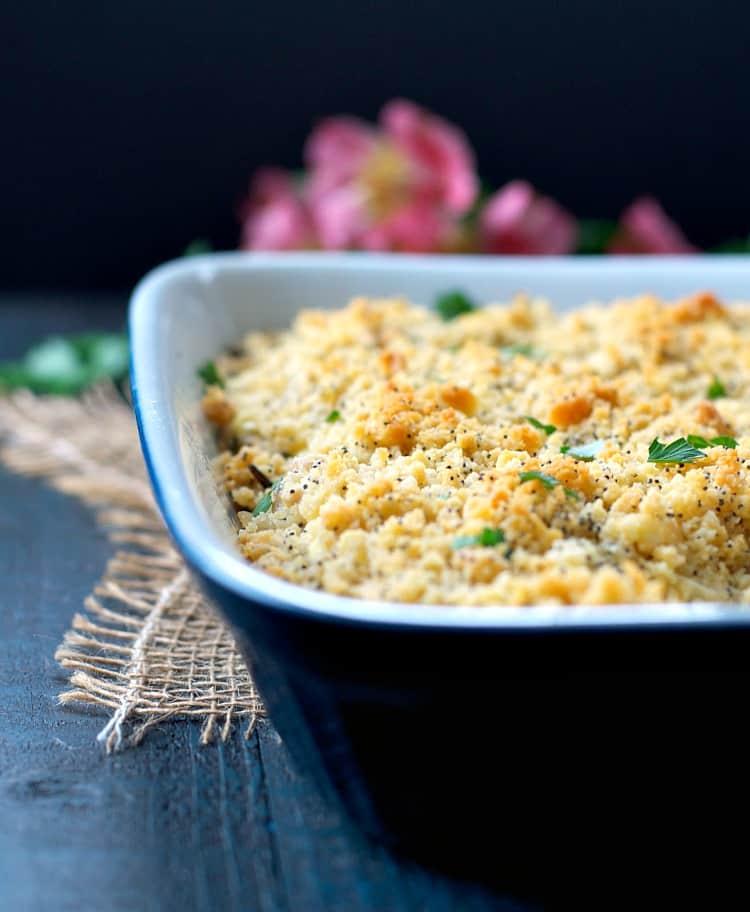 A close-up of a rectangular casserole dish filled with a golden-brown baked dish topped with breadcrumbs and herbs. The dish sits on a textured cloth, with pink flowers blurred in the background.