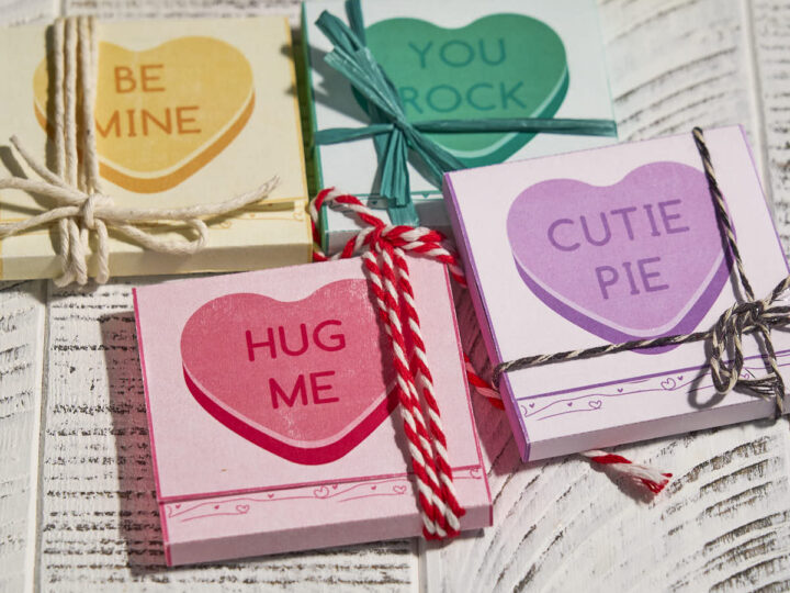 Four small packages of heart-shaped sticky notes on a wooden surface. Each package has a different colored heart with phrases: BE MINE, YOU ROCK, HUG ME, and CUTIE PIE. They are tied with colorful strings.