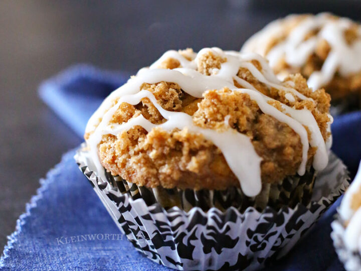 A close-up of a muffin topped with white icing and crumbly streusel, sitting in a black and white patterned cupcake liner on a blue cloth.