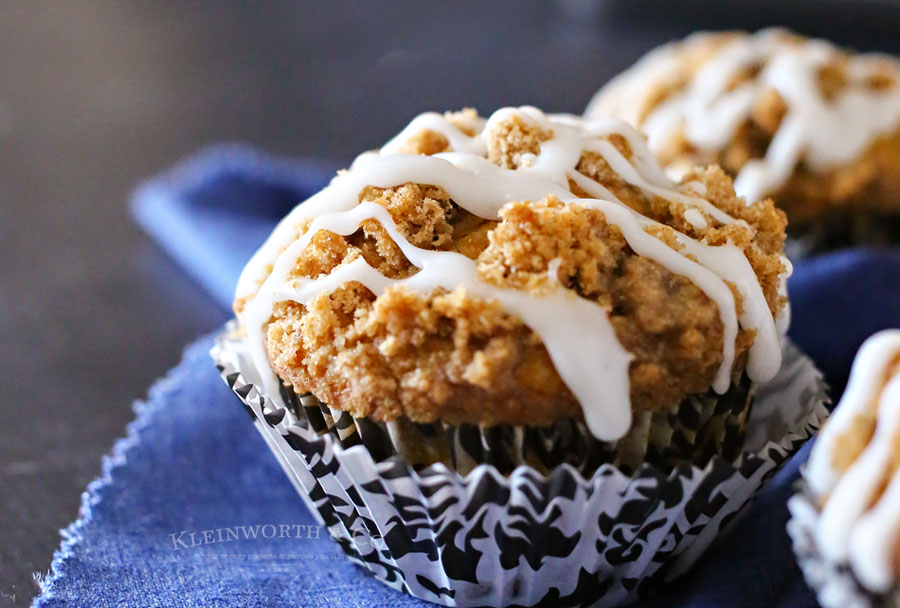 A close-up of a muffin topped with white icing and crumbly streusel, sitting in a black and white patterned cupcake liner on a blue cloth.
