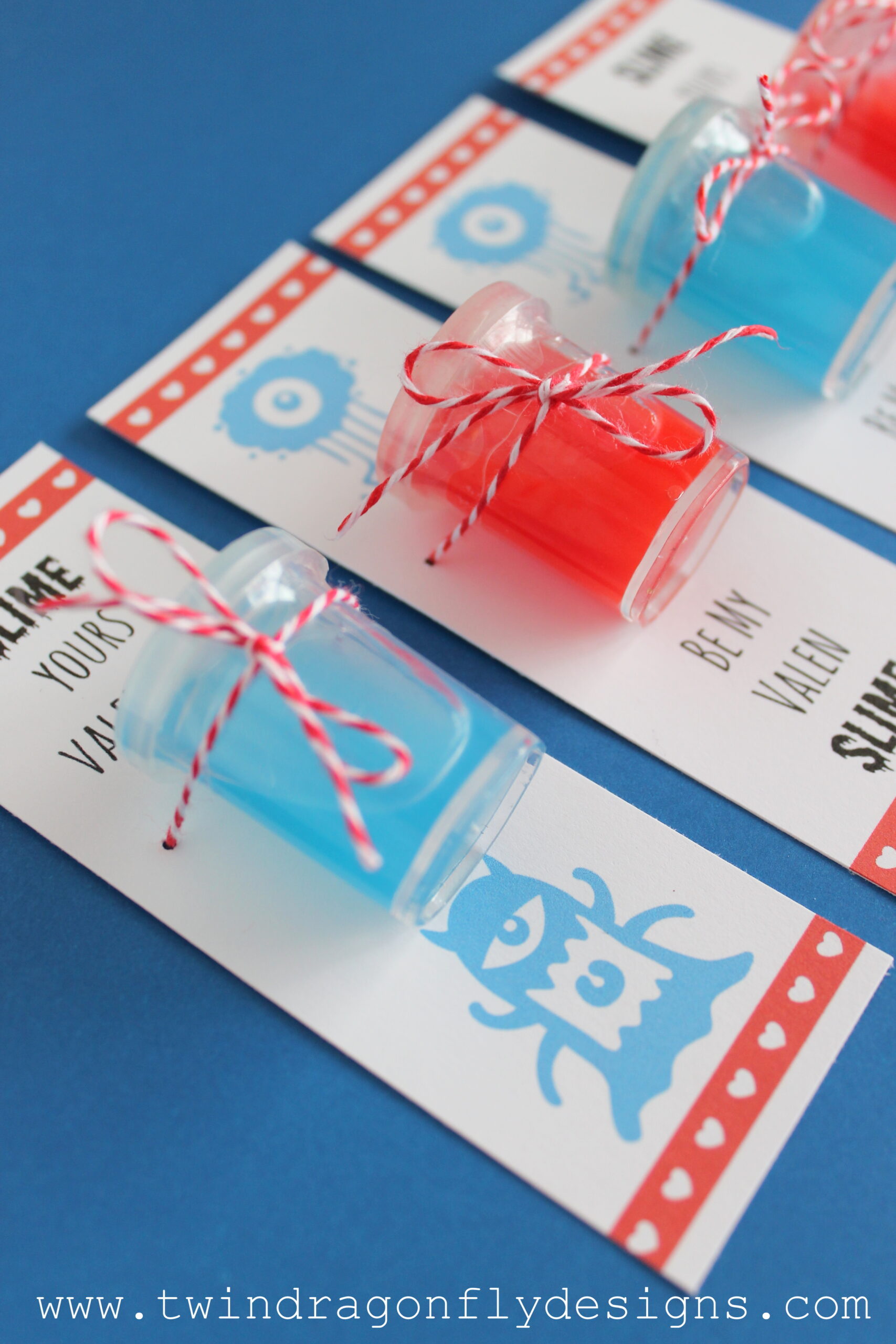 Three small jars with red and blue slime are attached to Valentines Day cards with red and white twine. The cards feature cute monster illustrations and playful text. The bottom of the image shows the website www.twindragonflydesigns.com.
