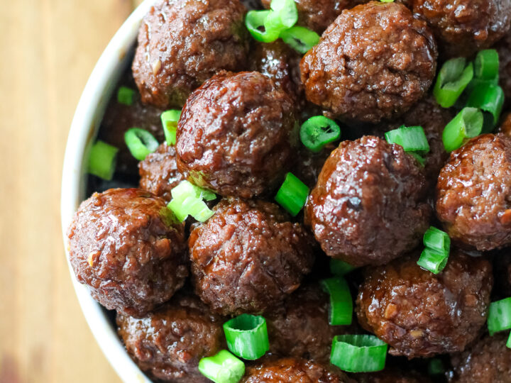 A bowl filled with glazed meatballs topped with chopped green onions is placed on a wooden surface. A single green onion is partially visible beside the bowl.