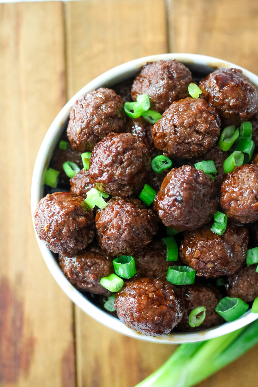 A bowl filled with glazed meatballs topped with chopped green onions is placed on a wooden surface. A single green onion is partially visible beside the bowl.