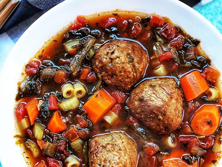 A bowl of hearty soup with three large meatballs, pasta, diced carrots, tomatoes, and greens in a rich broth. A striped cloth and a spoon sit nearby, along with a small bowl of grated cheese.