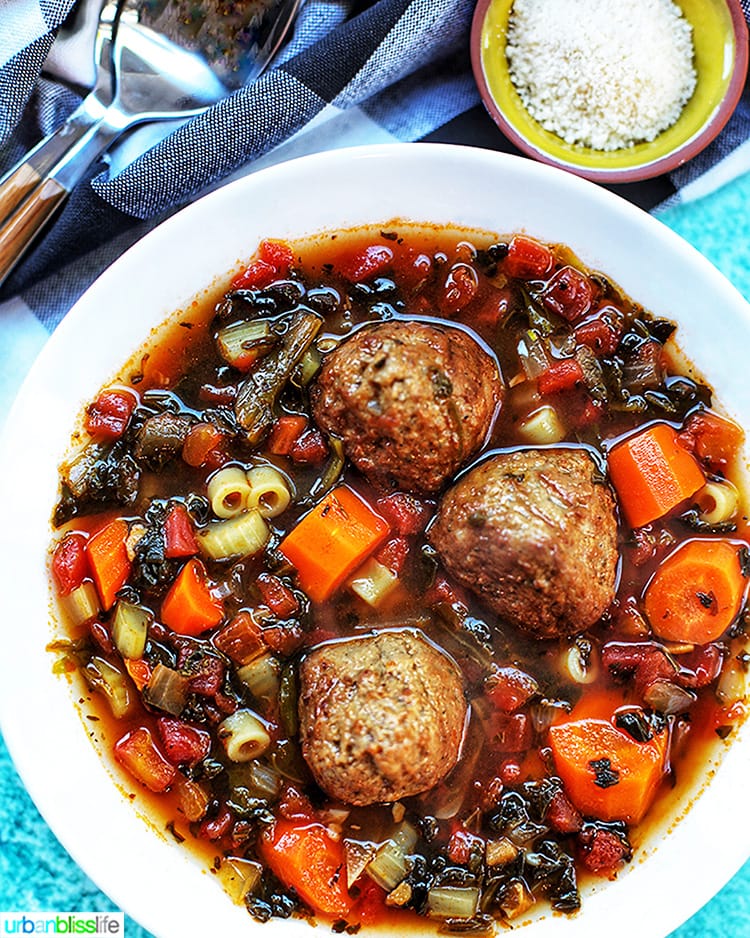 A bowl of hearty soup with three large meatballs, pasta, diced carrots, tomatoes, and greens in a rich broth. A striped cloth and a spoon sit nearby, along with a small bowl of grated cheese.