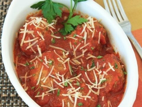 A white oval dish contains meatballs in marinara sauce, topped with grated cheese and garnished with parsley. The dish is placed on a woven placemat with a fork and a glass of wine nearby. Text at the bottom reads Slow Cooker Meatballs.