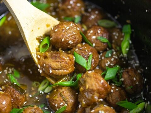 A wooden spoon scoops glazed meatballs from a dark pot. The meatballs are coated in a shiny sauce and garnished with sesame seeds and green onion slices.