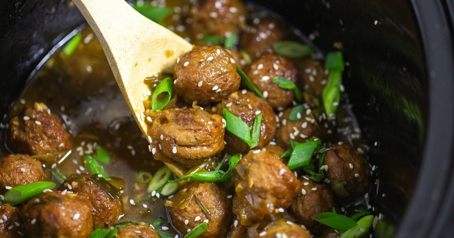 A wooden spoon scoops glazed meatballs from a dark pot. The meatballs are coated in a shiny sauce and garnished with sesame seeds and green onion slices.