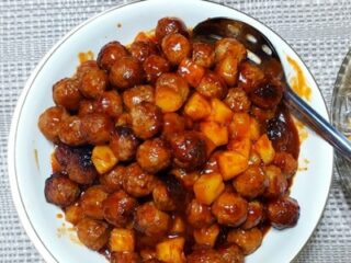 A bowl filled with saucy meatballs and diced pineapples, garnished with glaze. A slotted spoon rests in the bowl. On the side is a partial view of a plate with crescent rolls. The background features a woven gray and white placemat.