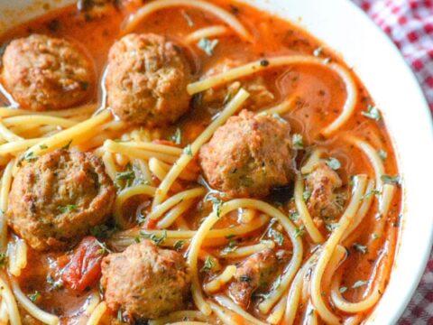 A white bowl filled with spaghetti and meatballs in a rich tomato soup. The dish is garnished with herbs. A piece of crusty bread is visible in the background on a red and white checkered cloth.