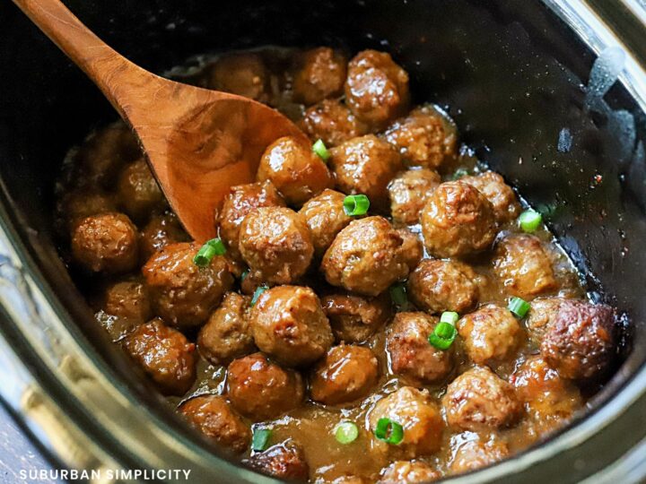 Slow cooker filled with glazed meatballs topped with chopped green onions. A wooden spoon is stirring the meatballs, which are in a rich, savory sauce. The image conveys a hearty and delicious dish.