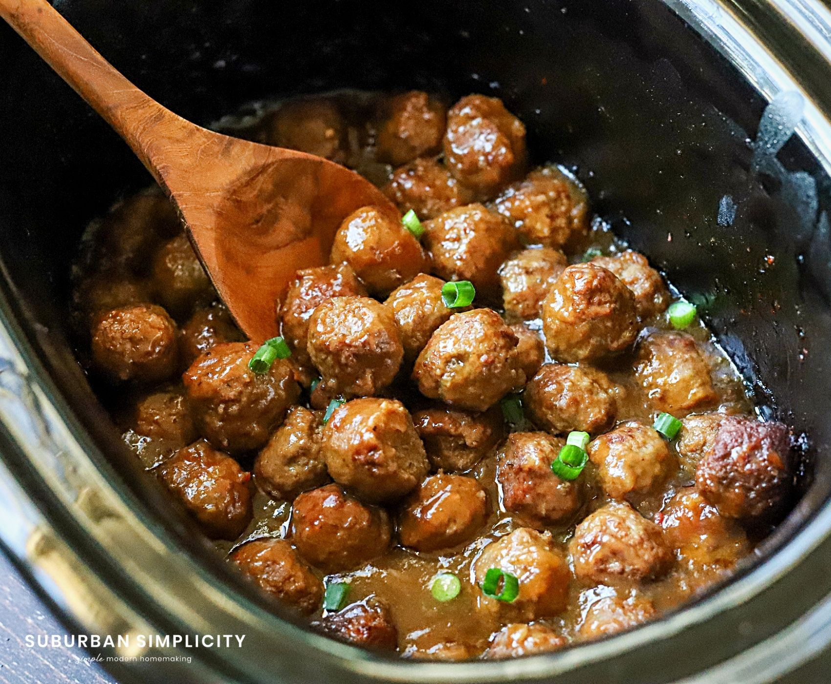 Slow cooker filled with glazed meatballs topped with chopped green onions. A wooden spoon is stirring the meatballs, which are in a rich, savory sauce. The image conveys a hearty and delicious dish.
