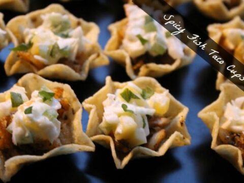 Close-up of spicy fish taco cups arranged on a dark surface. Each cup features a crispy shell filled with fish, topped with white sauce, diced green herbs, and a hint of spice. A diagonal text overlay reads Spicy Fish Taco Cups.
