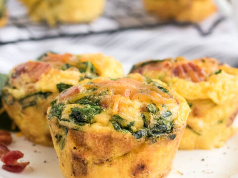 Three savory egg muffins with spinach and bits of bacon on a white plate. More muffins are visible in the background on a wire rack. The setting is a light kitchen countertop.