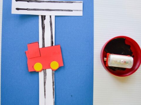 A craft project featuring a red truck glued onto a blue background. The truck is on a white paper road with black lines printed using a toy car dipped in paint. A toy car and paint dish are on the side.