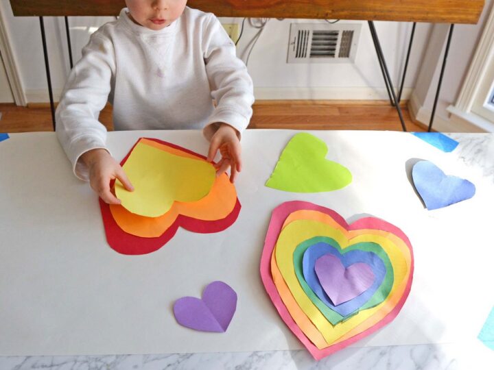 A child sits at a table with various colorful paper hearts in front of them. They are arranging layered hearts in rainbow colors, with individual purple, green, and yellow hearts scattered around. A brown table and toaster oven are in the background.