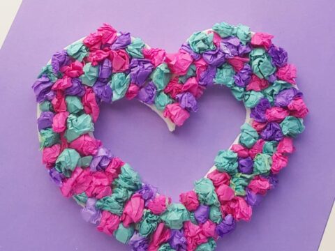 A heart-shaped wreath crafted from crumpled tissue paper in vibrant shades of pink, purple, turquoise, and green rests on a purple background. Perfect for adding a festive touch to your Valentines Day décor or exploring creative paper crafts.