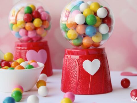 Two red gumball machines with heart designs, filled with colorful gumballs, and a white bowl of gumballs nearby. Pink background with heart shapes.