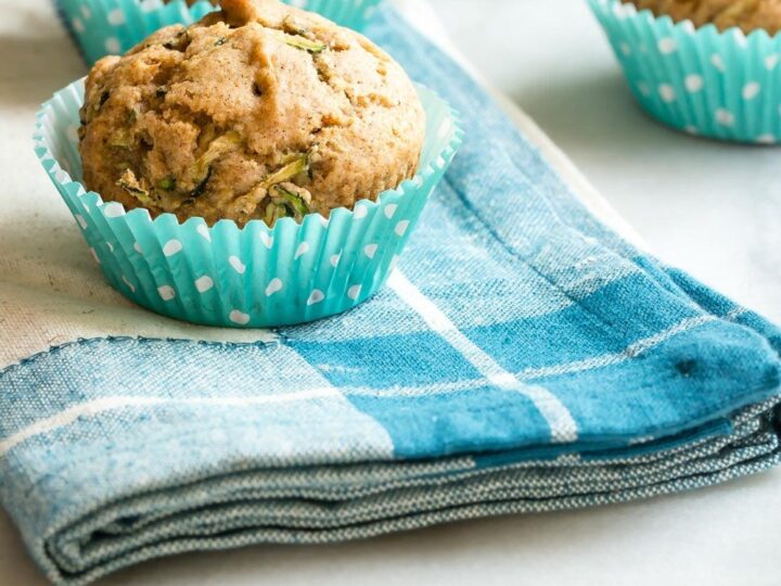 Four muffins in blue polka-dot paper cups are placed on a blue and white checkered cloth over a marble surface. The muffins appear freshly baked and are likely filled with nuts or seeds.