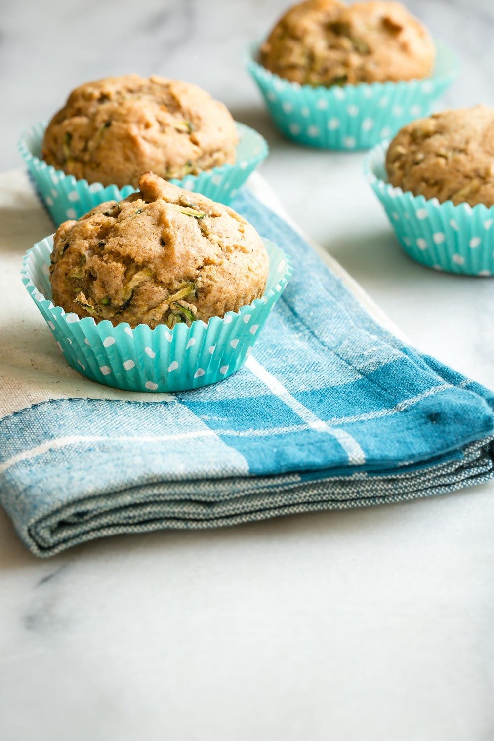 Four muffins in blue polka-dot paper cups are placed on a blue and white checkered cloth over a marble surface. The muffins appear freshly baked and are likely filled with nuts or seeds.