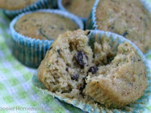 Close-up of zucchini muffins with a partially eaten one revealing a soft texture and chocolate chips inside. The muffins are in blue cupcake liners, resting on a green and white checkered cloth.