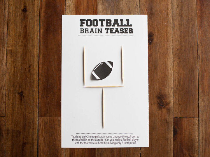 A brain teaser card on a wooden table features a goalpost made of toothpicks and a cutout of a football. The challenge text asks if you can rearrange the goalpost so the football is outside by moving only 2 toothpicks.