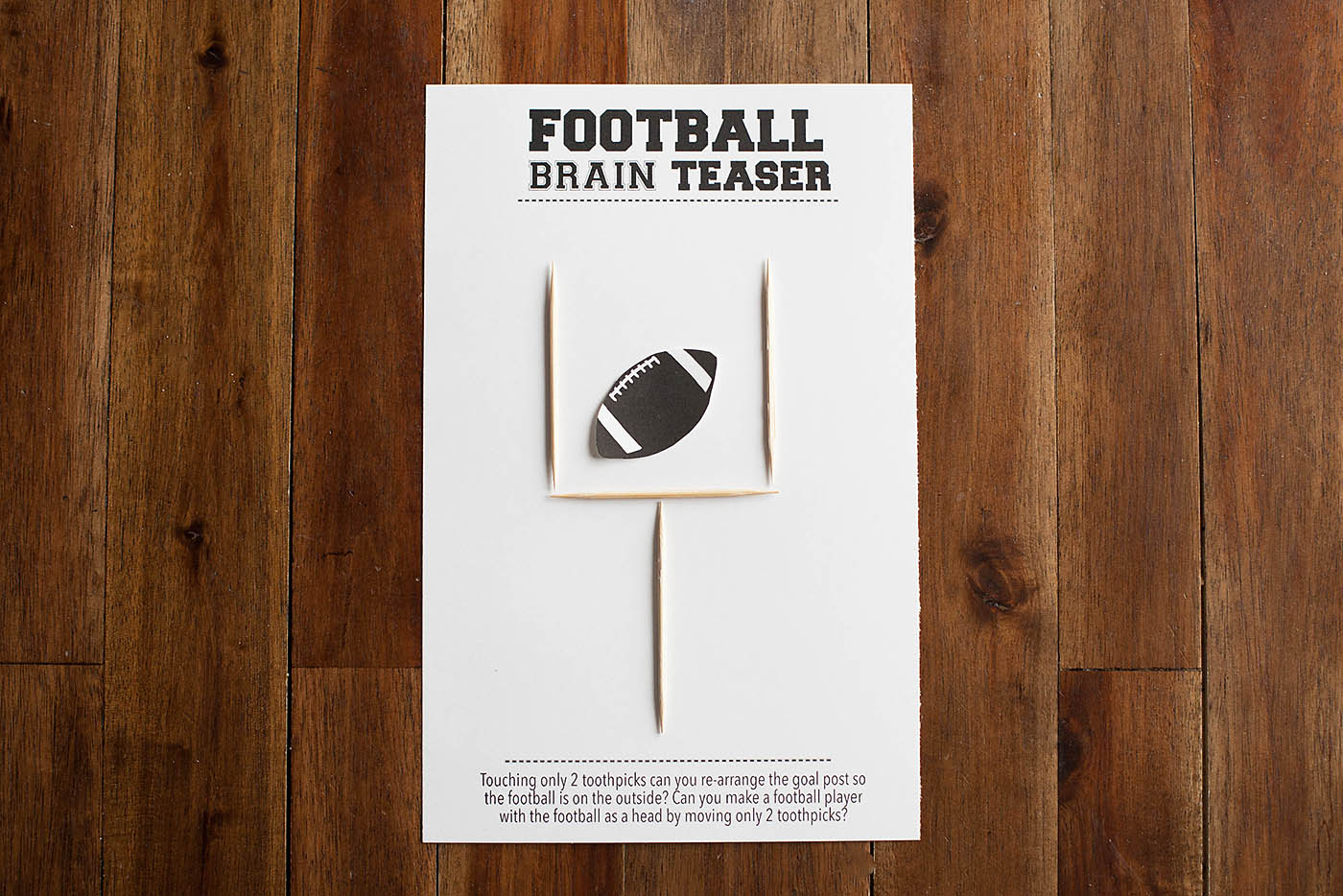 A brain teaser card on a wooden table features a goalpost made of toothpicks and a cutout of a football. The challenge text asks if you can rearrange the goalpost so the football is outside by moving only 2 toothpicks.