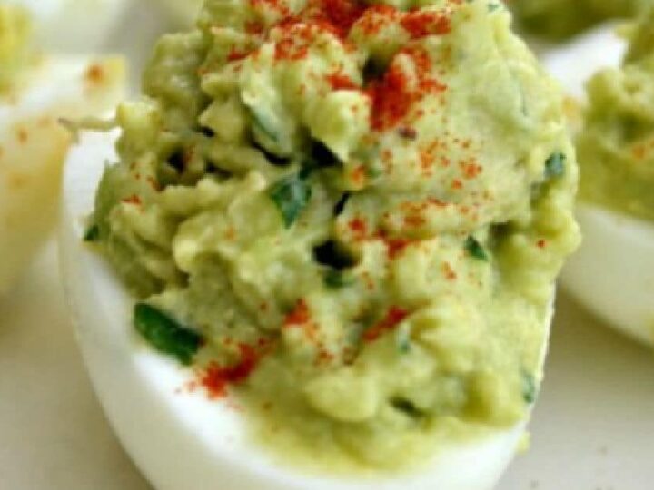 Close-up of deviled eggs topped with a green avocado mixture, garnished with a sprinkle of red paprika. The eggs are halved, with the filling piled high, displayed on a light-colored surface.