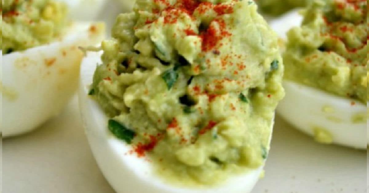 Close-up of deviled eggs topped with a green avocado mixture, garnished with a sprinkle of red paprika. The eggs are halved, with the filling piled high, displayed on a light-colored surface.
