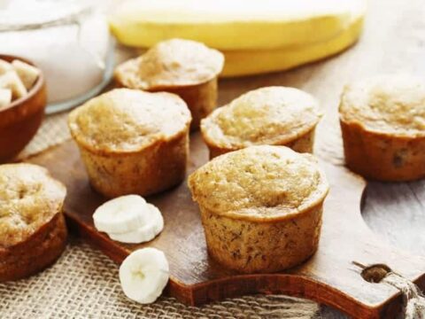 A wooden board with six freshly baked banana muffins. Slices of banana and a jar of sugar are in the background, adding to the cozy, homemade feel. The muffins are golden brown and appear moist and delicious.