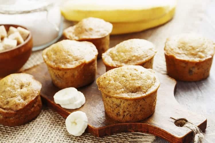 A wooden board with six freshly baked banana muffins. Slices of banana and a jar of sugar are in the background, adding to the cozy, homemade feel. The muffins are golden brown and appear moist and delicious.