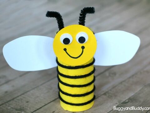 A colorful bee craft made from a yellow toilet paper roll with black pipe cleaner stripes and antennae. It has white paper wings and googly eyes, with a smiling face drawn on. The background is a wooden surface.