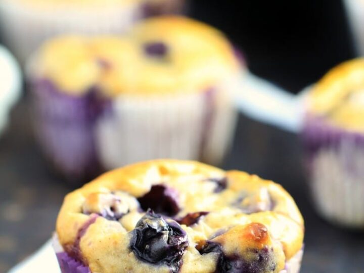 A close-up of a blueberry muffin in a white paper liner, showcasing vibrant blueberries baked into a golden, fluffy surface. In the background, more muffins are slightly blurred against a dark backdrop.