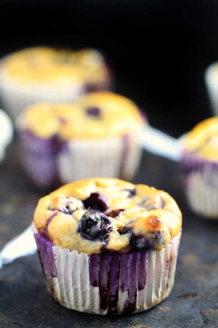 A close-up of a blueberry muffin in a white paper liner, showcasing vibrant blueberries baked into a golden, fluffy surface. In the background, more muffins are slightly blurred against a dark backdrop.