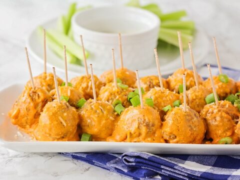 A platter of buffalo chicken meatballs topped with sliced green onions, each with a toothpick. The dish is on a blue and white checkered cloth. In the background, there is a bowl and celery sticks.