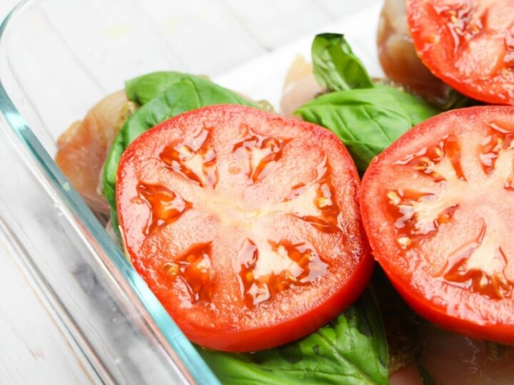 A glass baking dish with raw chicken breasts, fresh basil leaves, and sliced tomatoes on top. The dish is placed on a white wooden surface.