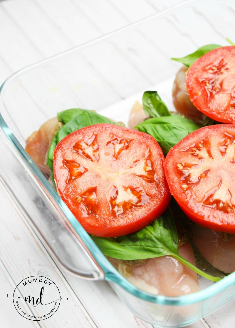 A glass baking dish with raw chicken breasts, fresh basil leaves, and sliced tomatoes on top. The dish is placed on a white wooden surface.