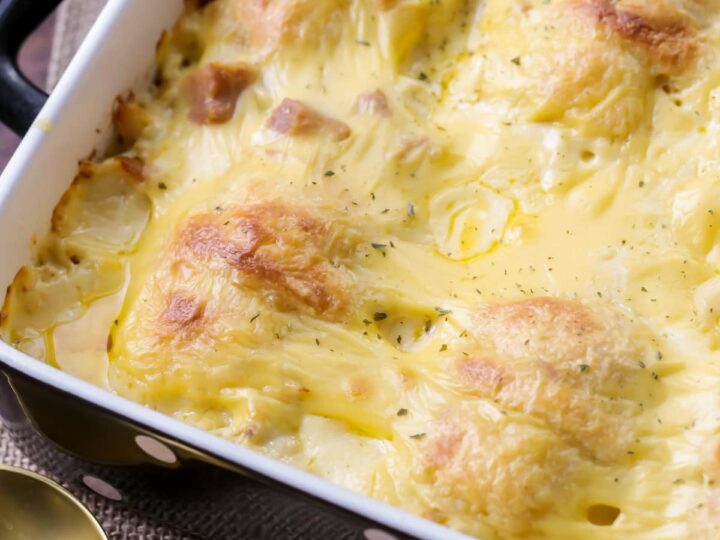 A baked potato dish in a rectangular pan, topped with golden, melted cheese. The surface appears creamy, with slightly browned edges. A gold spoon is placed beside the pan, resting on a woven surface.