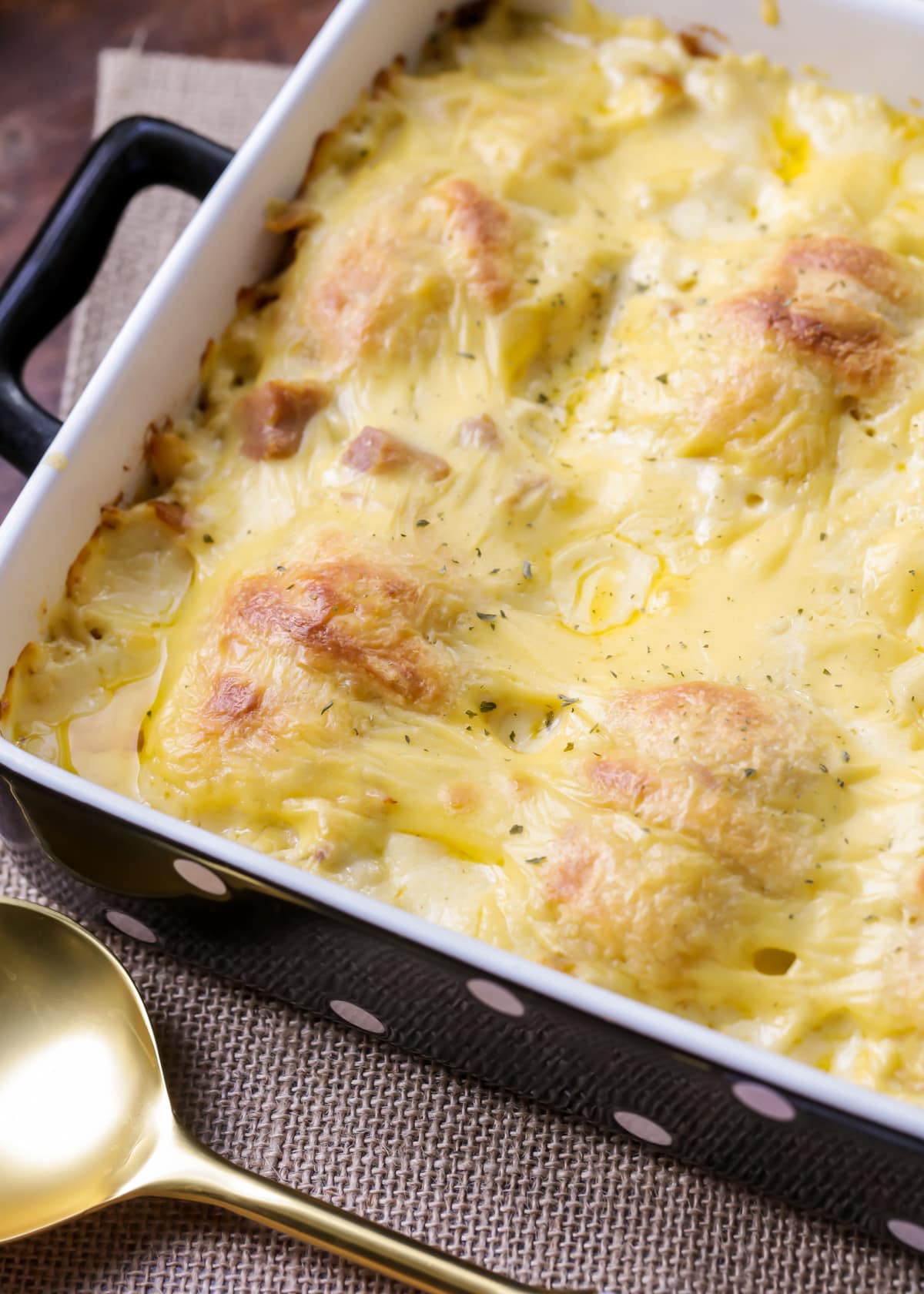 A baked potato dish in a rectangular pan, topped with golden, melted cheese. The surface appears creamy, with slightly browned edges. A gold spoon is placed beside the pan, resting on a woven surface.