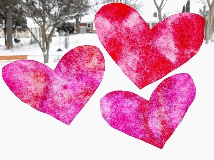 Three watercolor paper hearts in shades of red and pink are displayed on a window. Snowy trees and houses are visible in the blurred background, giving a wintry outdoor view.