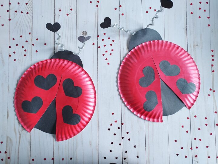 Two ladybug crafts made from red paper plates with black heart spots and black heads. Silver pipe cleaners form antennae. Black hearts and dots are scattered on a light wooden background.