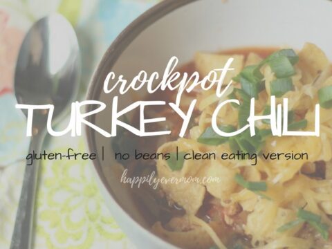 A bowl of crockpot turkey chili topped with shredded cheese and chopped herbs. Text overlay reads crockpot TURKEY CHILI with descriptors gluten-free, no beans, and clean eating version. A spoon and colorful napkin are beside the bowl.