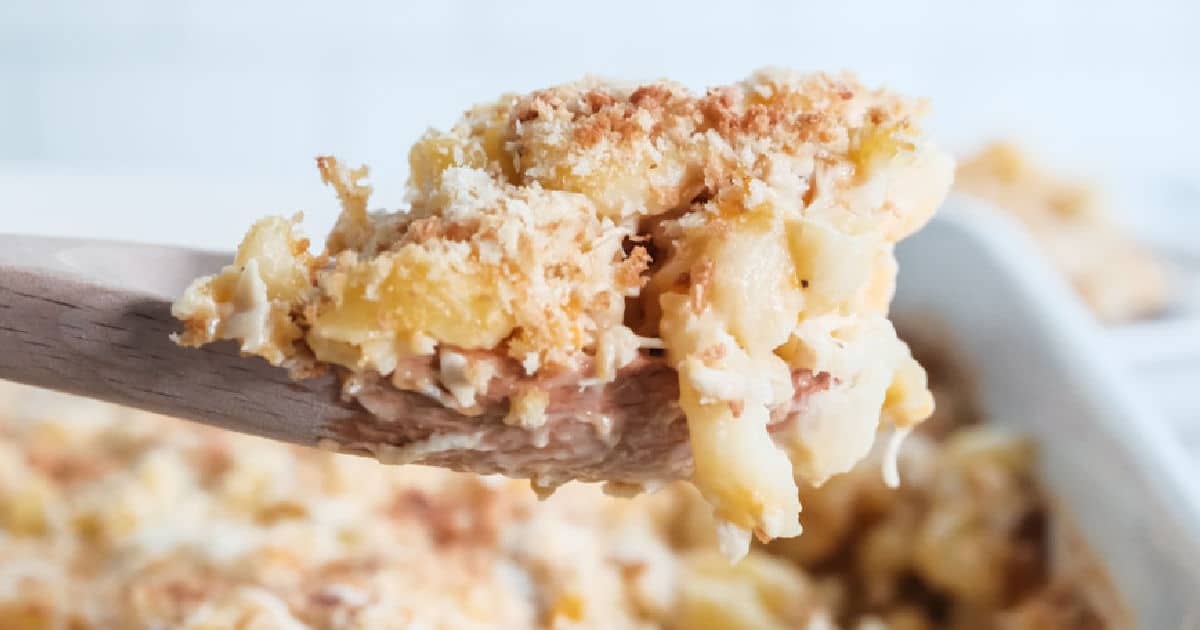 A wooden spoon holds a serving of creamy macaroni and cheese, topped with a golden breadcrumb crust. The dish is in the background, partially visible, showcasing the baked cheesy texture and crumb topping.