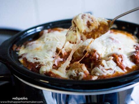 Cheese-covered meatballs being lifted from a slow cooker filled with a rich tomato sauce and melted cheese. The slow cooker is black with a glossy finish.