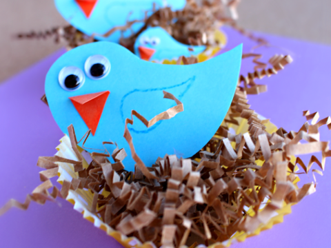 Three paper bluebirds with googly eyes and orange beaks sit in nests made from brown crinkled paper in yellow paper cups. The background is purple.