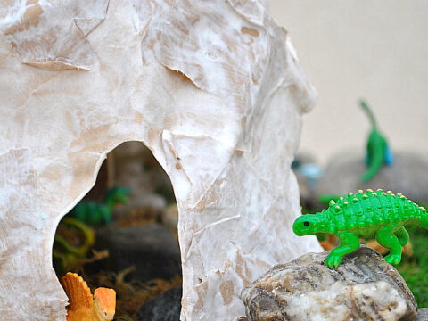 A paper mache cave is displayed on a bed of moss, with small toy dinosaurs, including a green one on a rock. The text reads Dinosaur Paper Mache Cave.