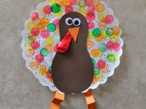 A colorful turkey craft made from a paper plate. The turkey has a brown body, large googly eyes, an orange beak, and red wattle. The tail features multicolored tissue paper circles. The legs are orange strips of paper.